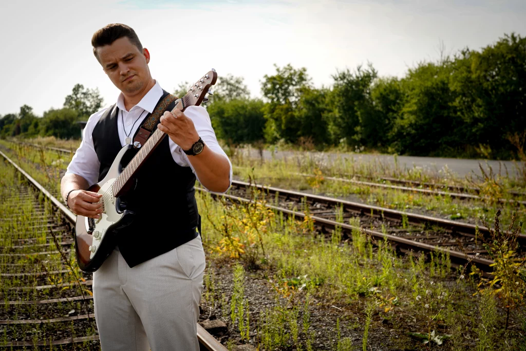 David Schneider Gitarre spielend an einem alten Bahnhof, auf den Gleisen.